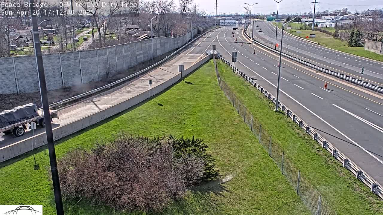 A mostly empty highway curves gently to the right, bordered by grassy areas and a chain-link fence under a sunny sky.