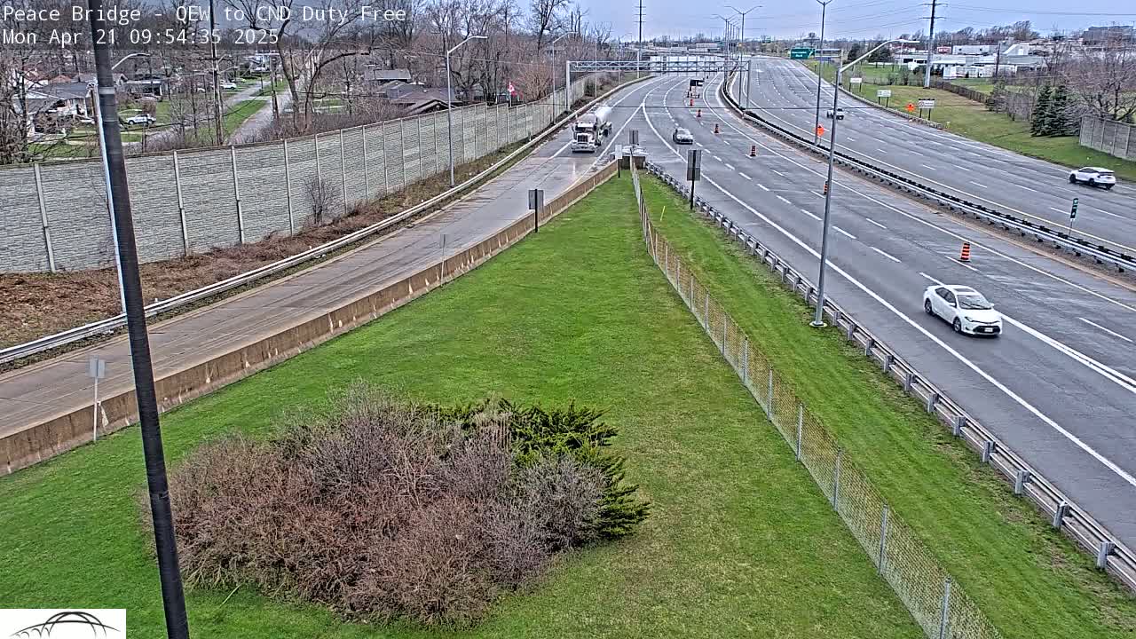 A multi-lane highway with a few cars and a tanker truck under overcast skies, bordered by grassy areas and a chain link fence.