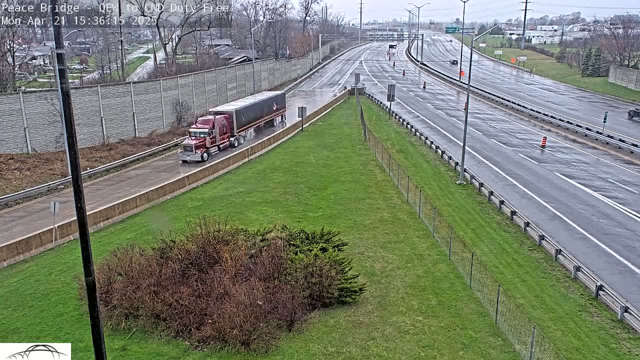 A red semi-truck travels on a wet highway next to a grassy area enclosed by a chain link fence, under an overcast sky.