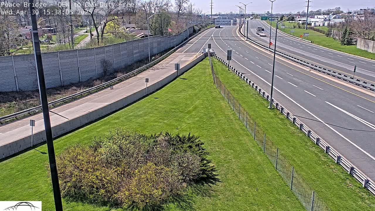 A highway scene on a sunny day shows two lanes of traffic, grassy areas separated by chain link fences, and a concrete wall bordering residential areas.