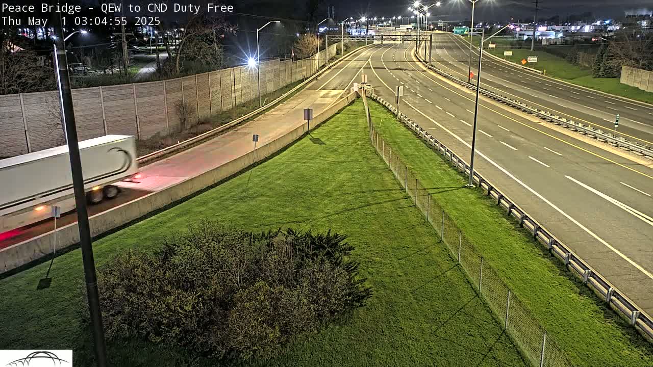 A white semi-truck drives on a highway at night, next to a grassy area bordered by a chain-link fence.
