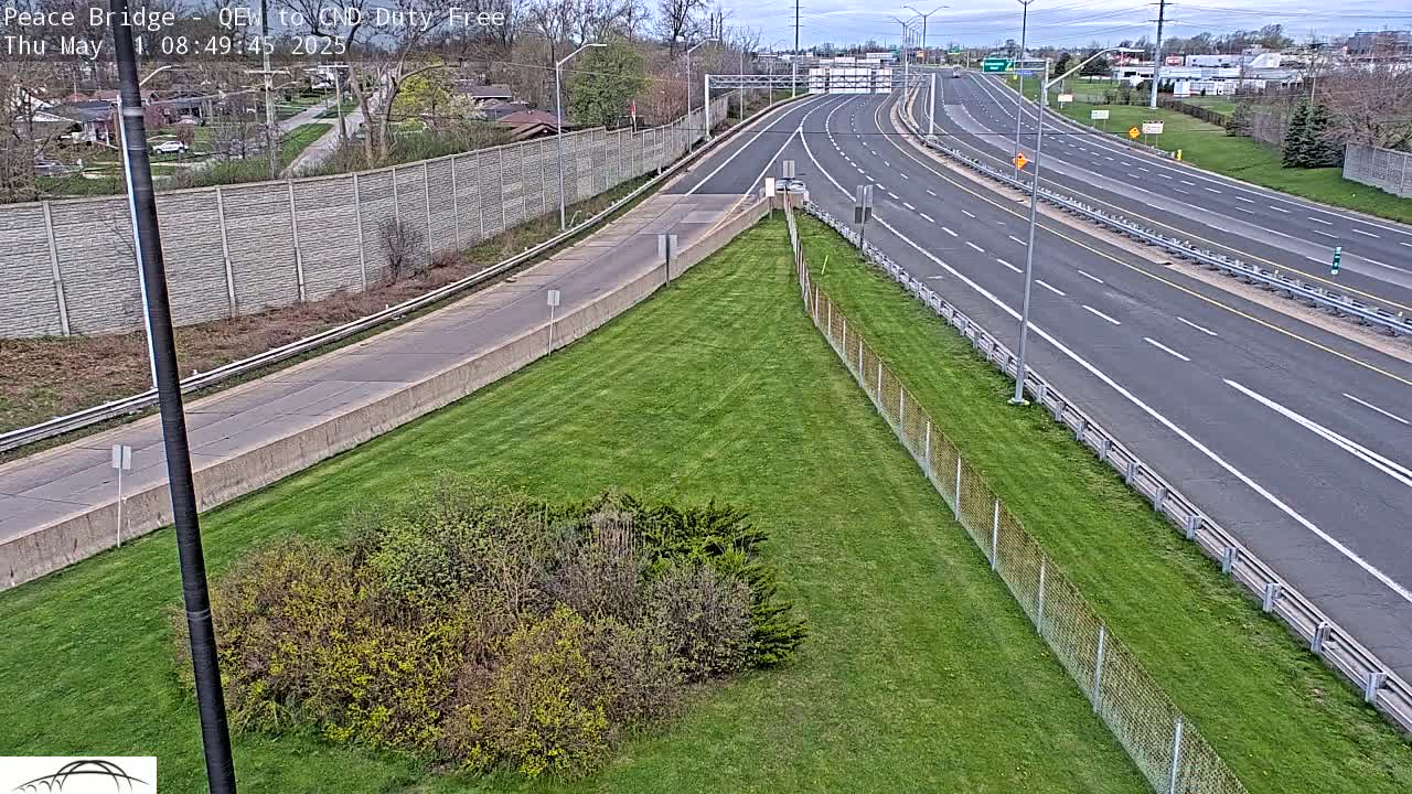 A mostly empty highway curves gently to the right, bordered by a grassy area with shrubs and a chain-link fence under a partly cloudy sky.