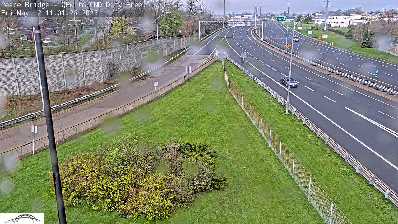 A rain-slicked highway with several cars driving on it, seen from an elevated vantage point overlooking a grassy area with a chain link fence.