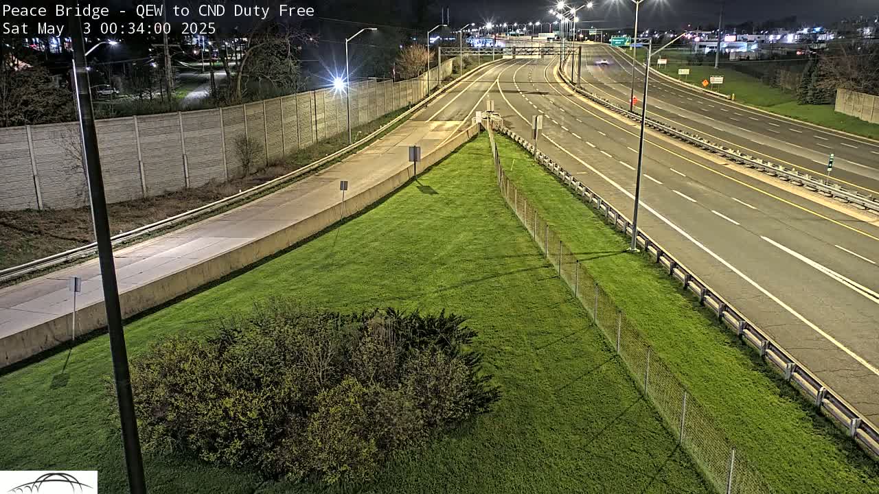 A nighttime aerial view shows a multi-lane highway with few vehicles, bordered by grassy areas and chain-link fencing.