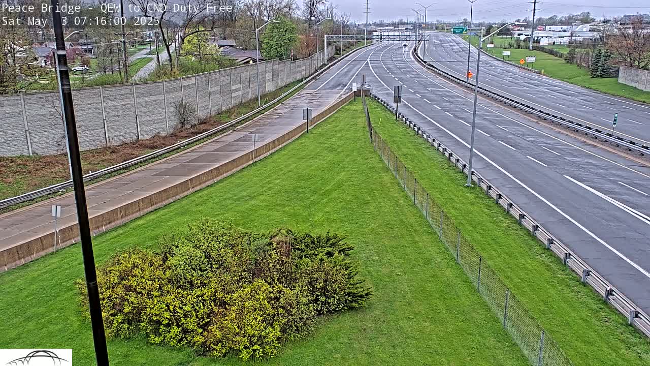 A wet, multi-lane highway stretches into the distance, bordered by green grass and a chain link fence, with a few cars visible in the far distance.
