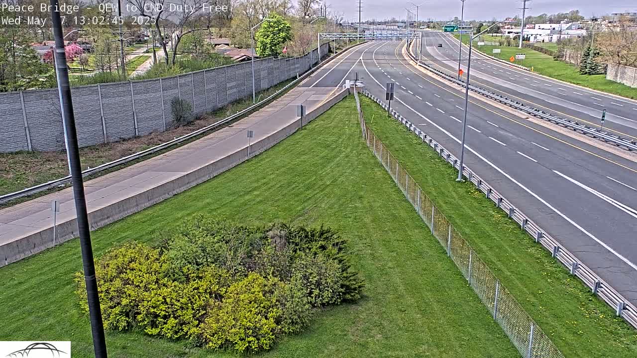 A highway with few cars is seen on an overcast day, bordered by grassy areas, shrubs, and a chain-link fence.