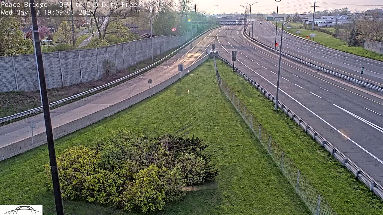 A highway scene at dusk shows a multi-lane roadway with sparse traffic, bordered by grassy areas and a chain-link fence.