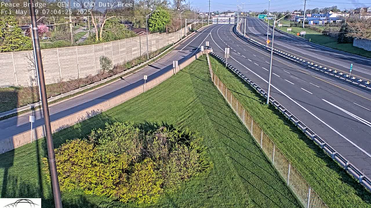 A highway with light traffic, bordered by grassy slopes and chain-link fences, under a sunny sky.