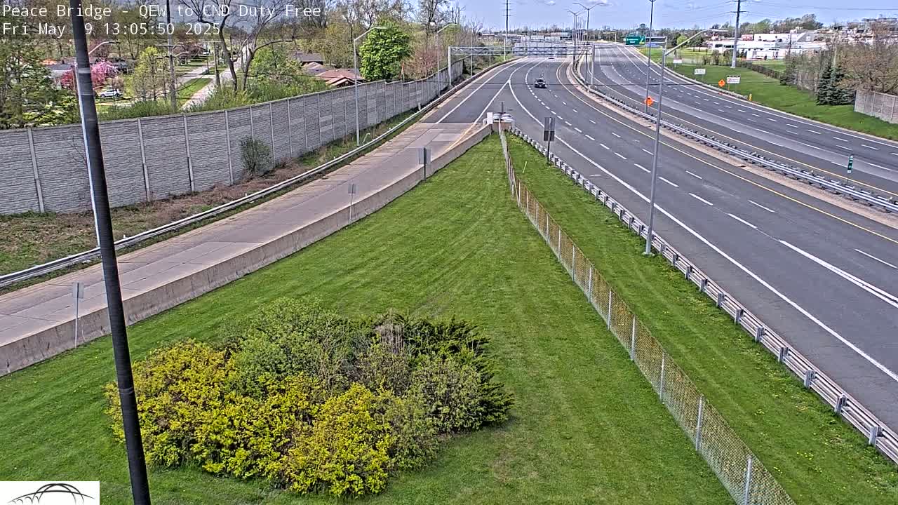 A highway scene on a sunny day shows a multi-lane highway curving to the right, with green grass and shrubbery along the side, bordered by a chain-link fence.