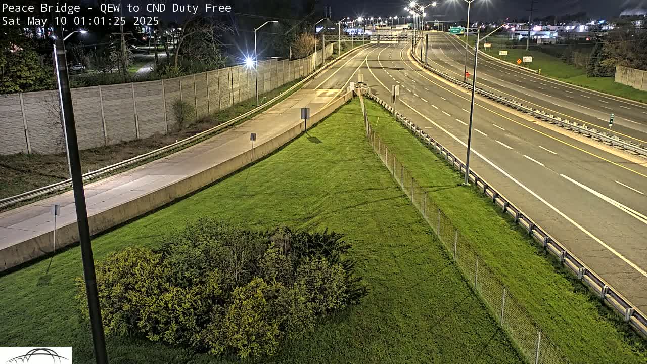 A nighttime, high-angle view shows a multi-lane highway with sparse traffic, bordered by grassy areas and a chain-link fence.