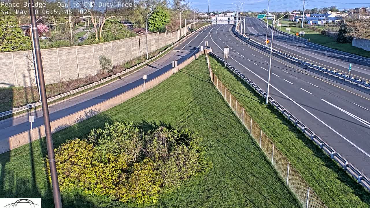A highway with sparse traffic on a sunny day, bordered by grassy areas and a chain-link fence.