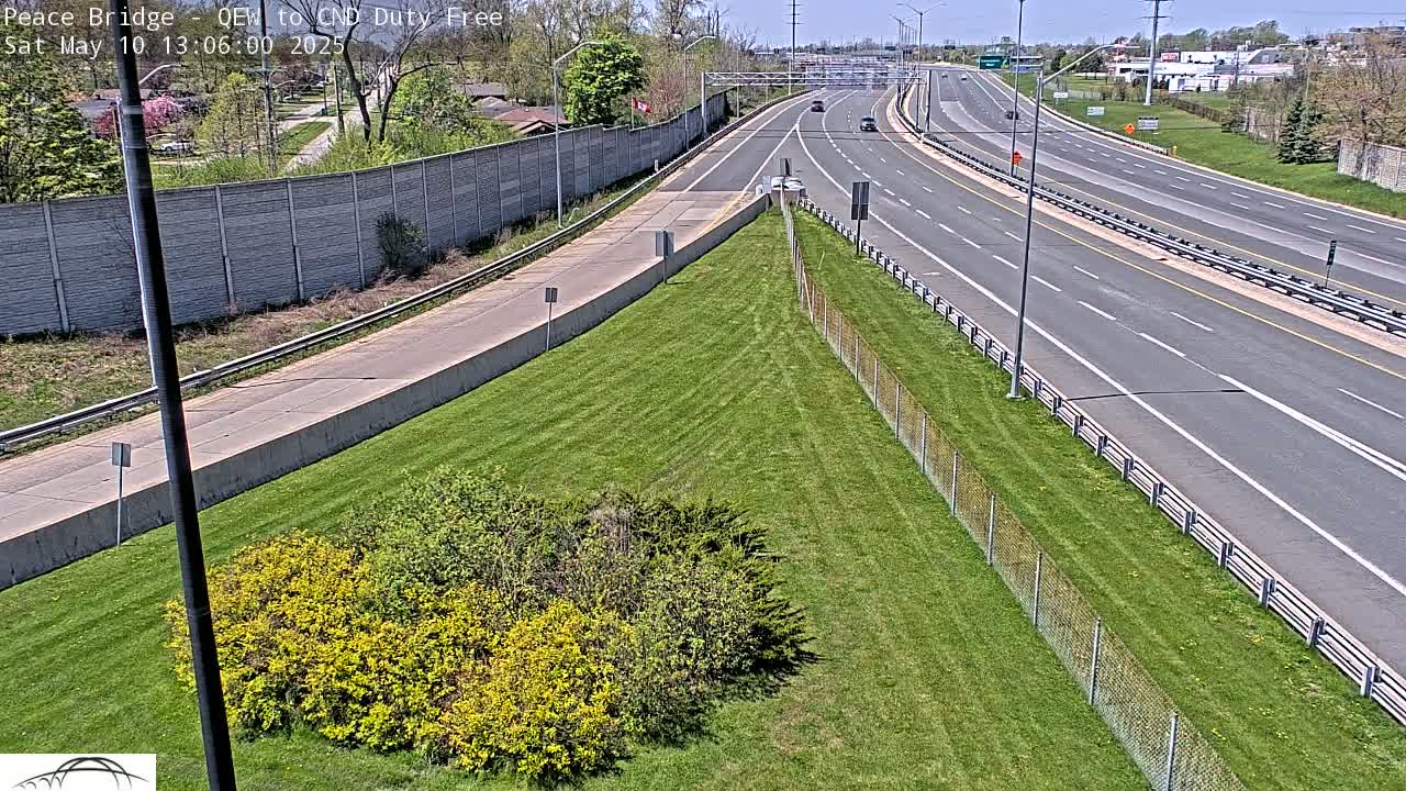 A sunny day reveals a highway scene with several vehicles, bordered by a grassy area with shrubs and a chain-link fence.