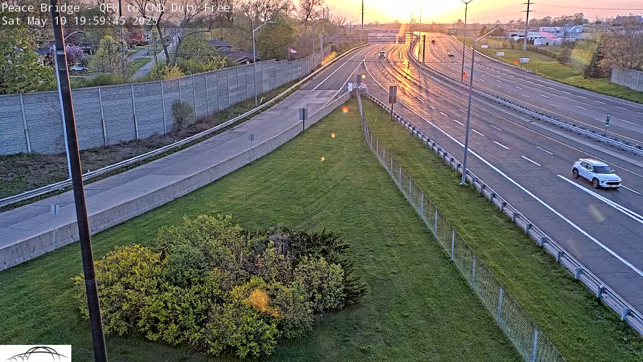 A multi-lane highway at sunset with a few cars, bordered by grassy areas and a chain-link fence.