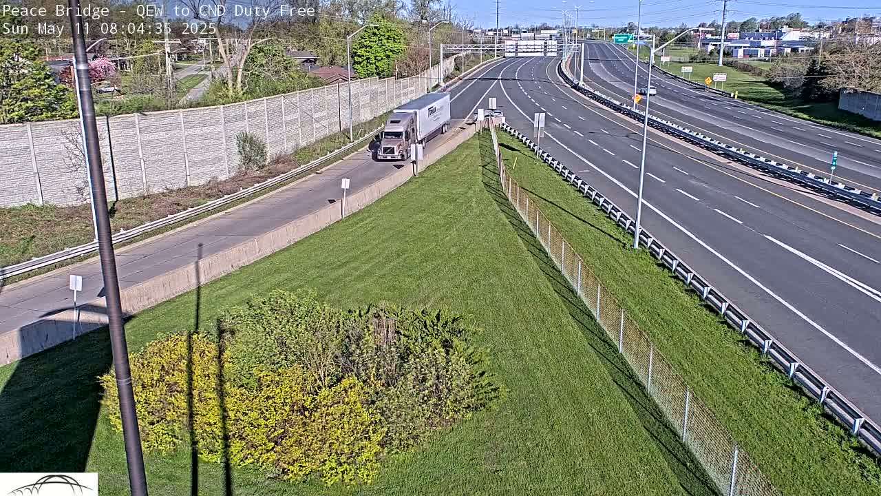 A semi-truck is on an off-ramp of a multi-lane highway on a sunny day with green grass and shrubs along the side.