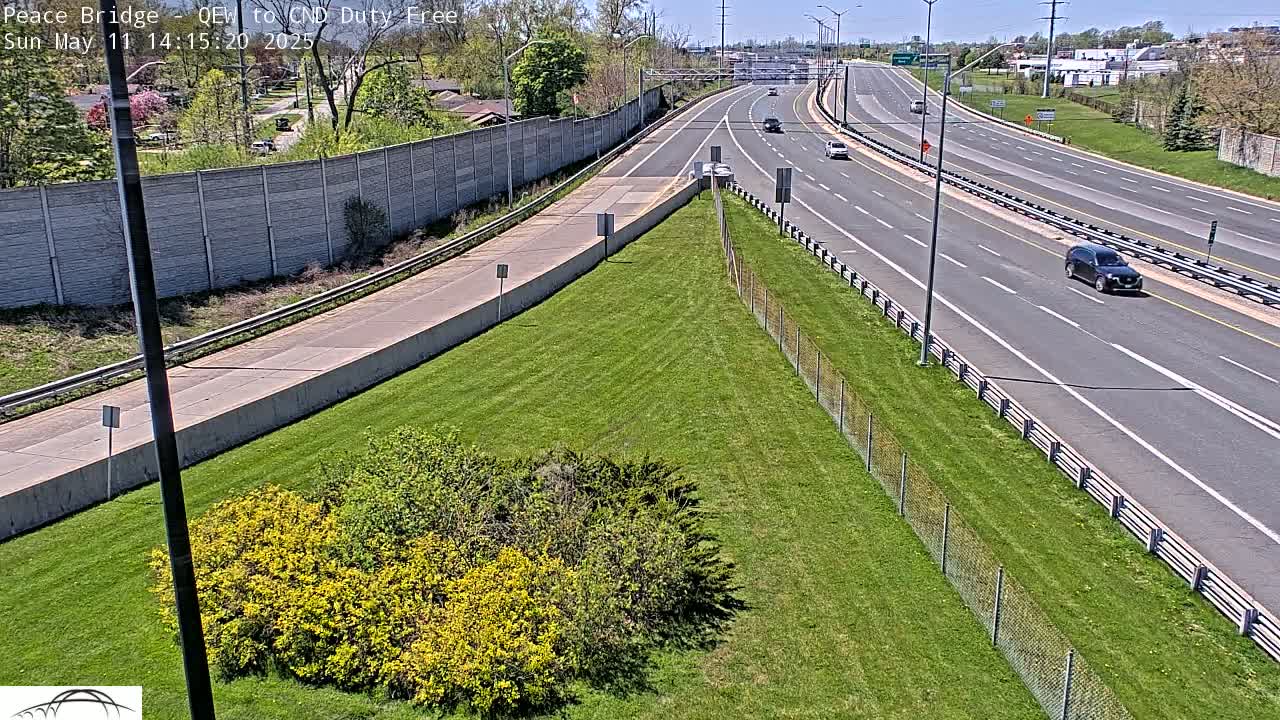 A highway on a sunny day with several cars driving, bordered by green grass, shrubs, and a concrete wall.