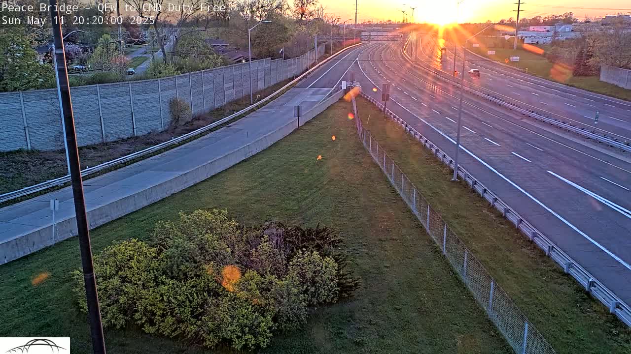 A multi-lane highway with sparse traffic is seen at sunset, bordered by grassy areas and a chain-link fence.