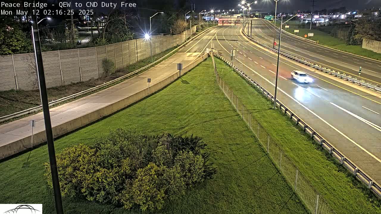 A nighttime view of a highway with sparse traffic, bordered by grassy areas and a fence, under clear skies.