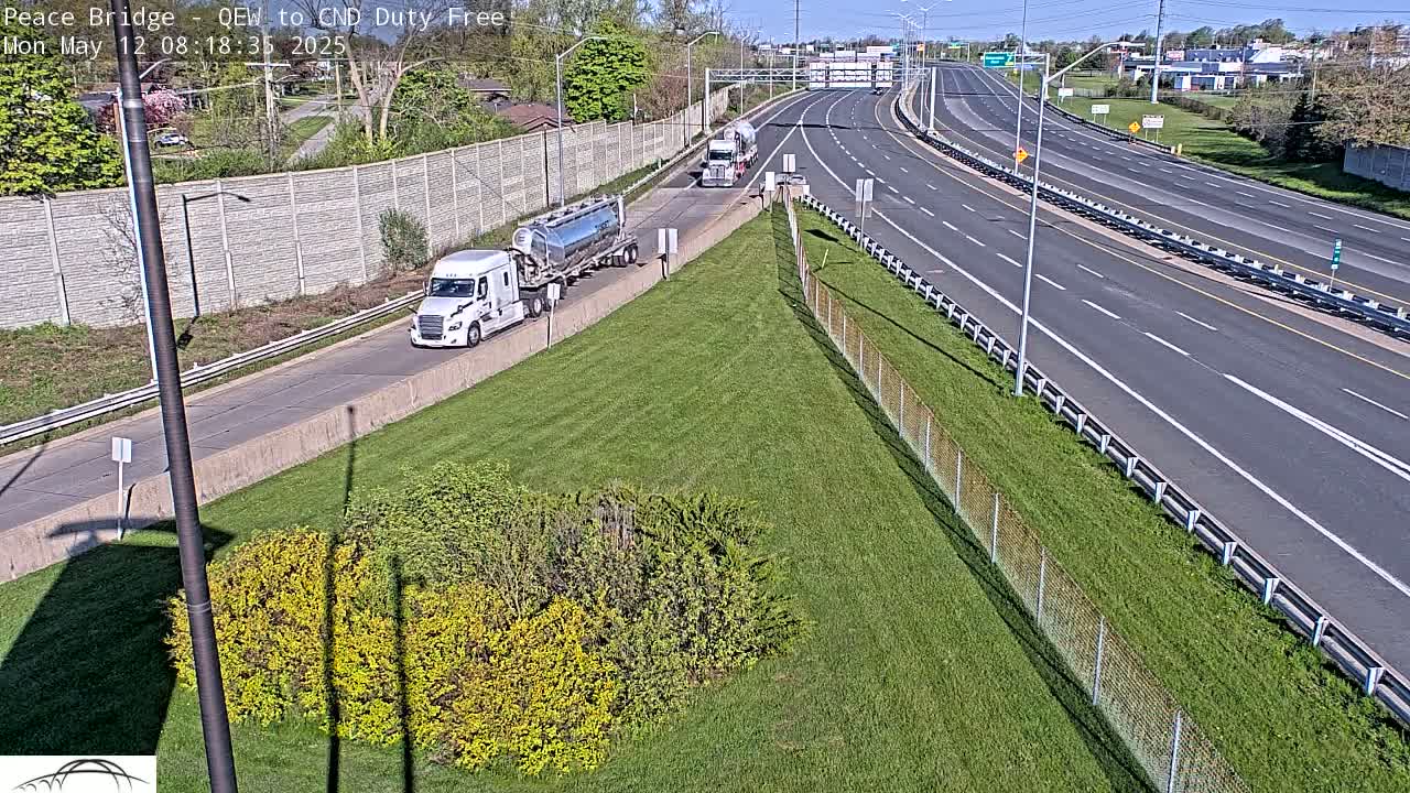 Two semi-trucks are parked on the side of a multi-lane highway on a sunny day, with green grass and shrubs visible nearby.