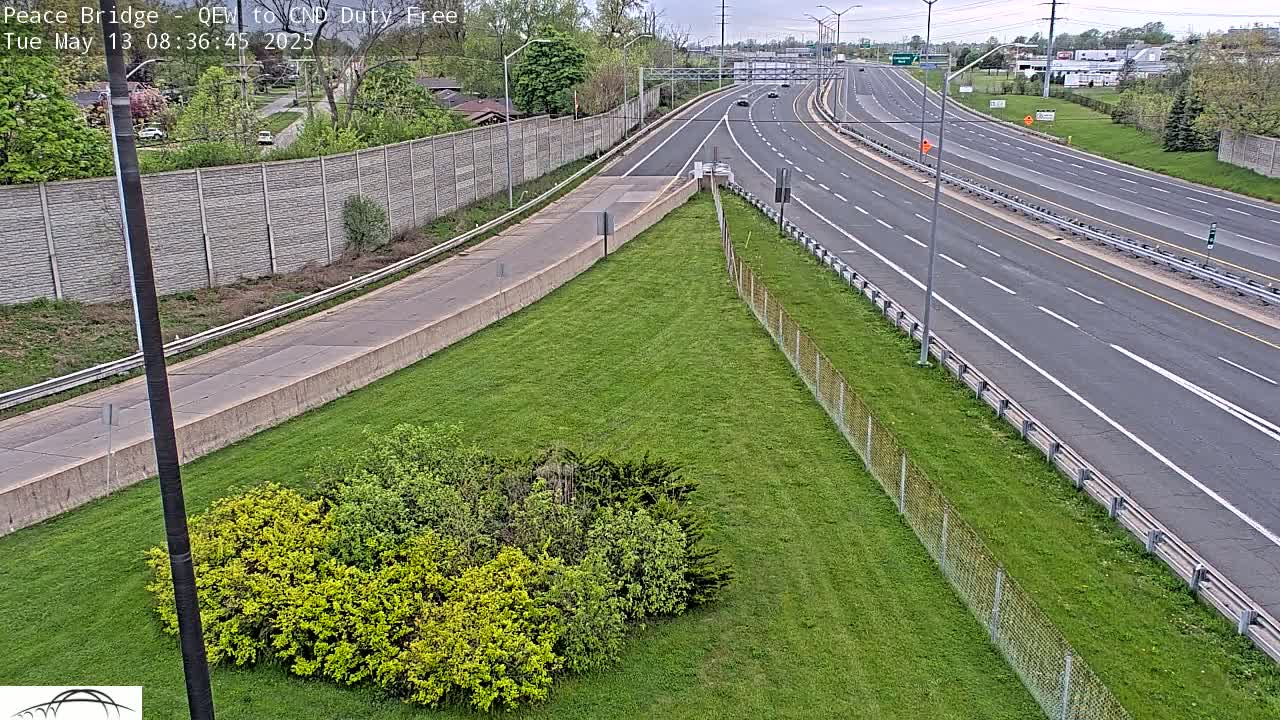 A mostly empty highway with a few cars visible under a partly cloudy sky, bordered by grassy areas and a chain-link fence.