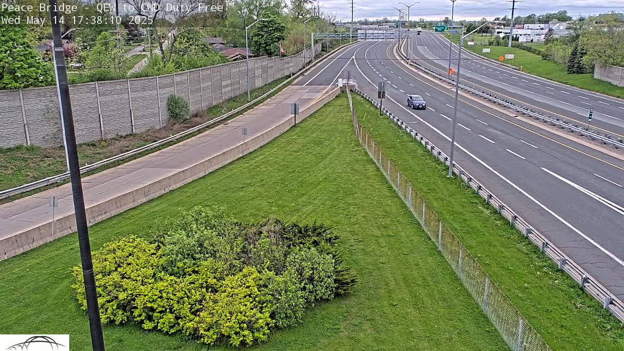 A grassy area with a small grouping of shrubs sits beside a multi-lane highway under a partly cloudy sky.