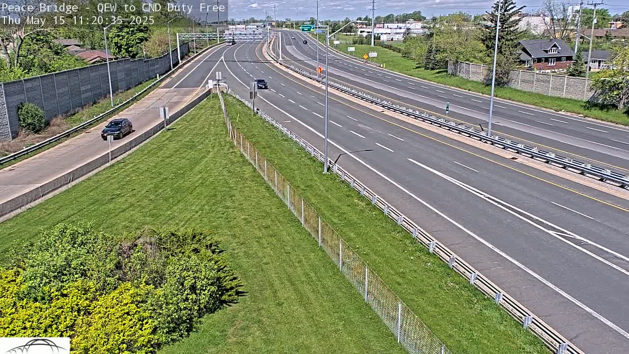A mostly empty highway stretches into the distance under a partly cloudy sky, bordered by grassy areas and chain link fences.
