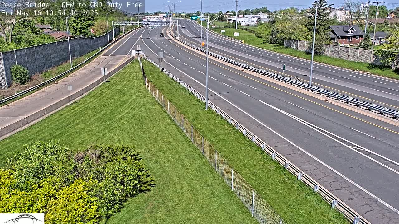 A multi-lane highway with light traffic on a sunny day, bordered by grassy areas and chain-link fencing.