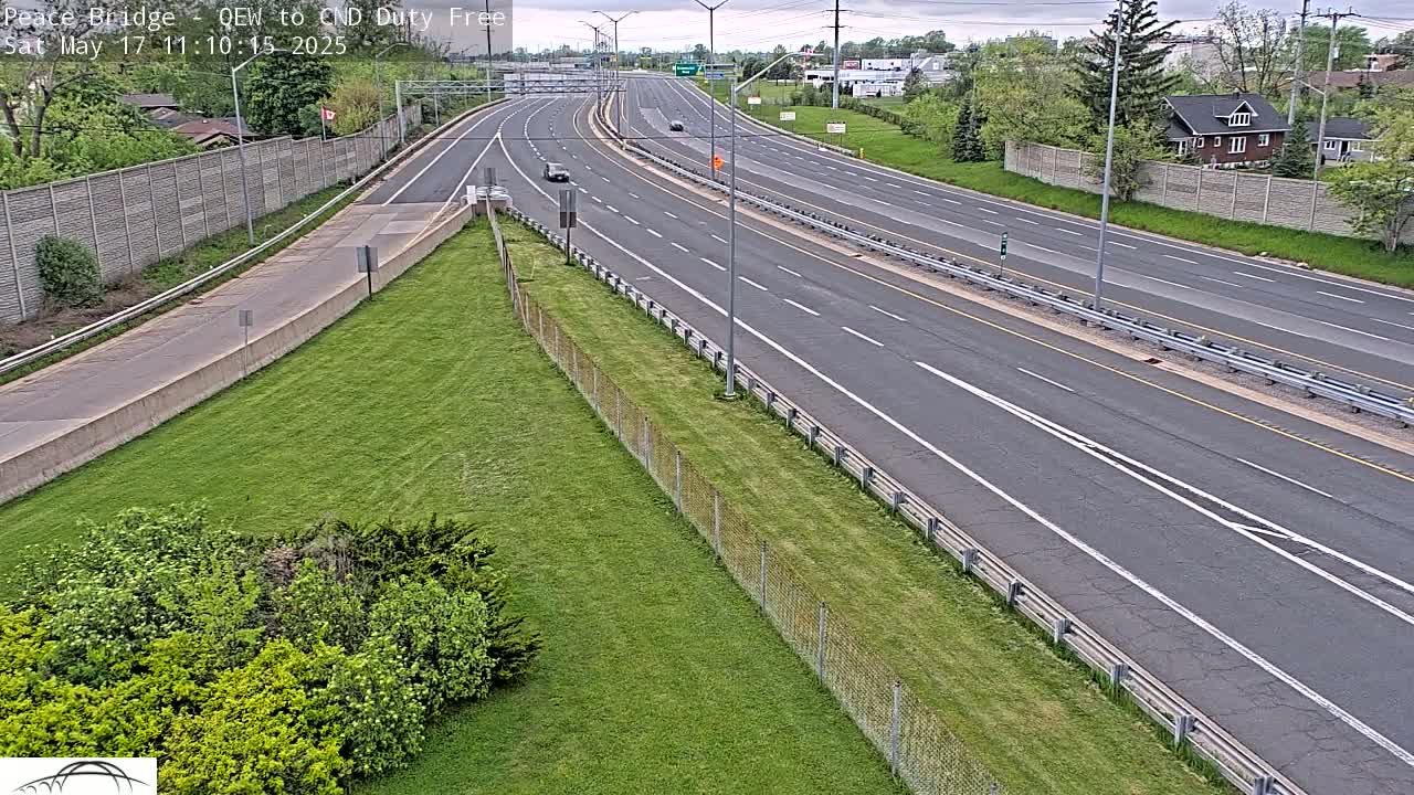 A multi-lane highway with sparse traffic stretches into the distance under a partly cloudy sky, bordered by grassy areas and chain link fencing.
