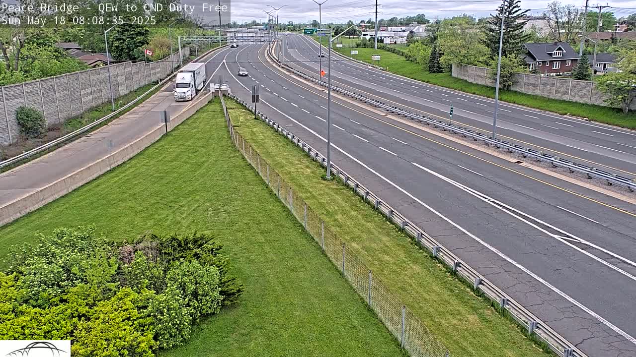 A multi-lane highway with several vehicles driving on it under a partly cloudy sky, bordered by grassy areas and chain link fences.