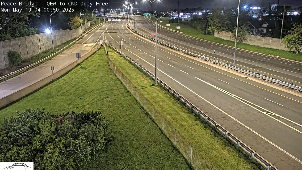 A nighttime, high-angle view of a multi-lane highway with sparse traffic, bordered by grassy areas and fences.