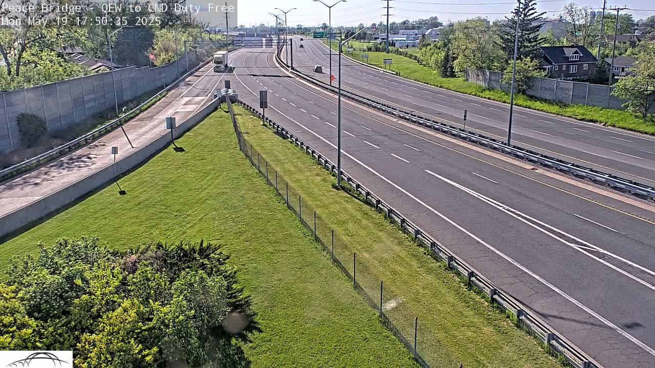 A sunny day shows a multi-lane highway with sparse traffic, bordered by grassy areas and a chain-link fence, with a building visible in the distance.