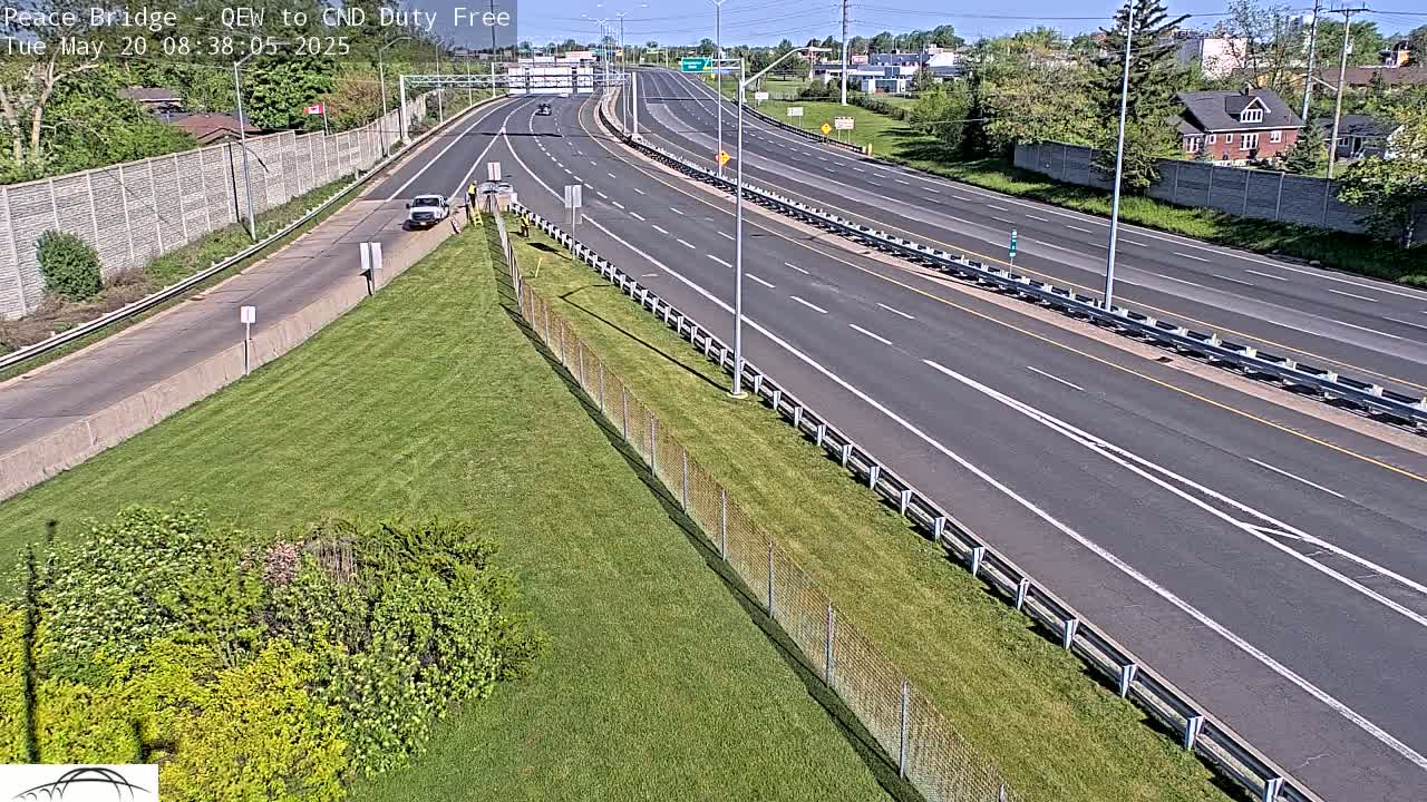 A highway with a few cars on a sunny day, bordered by grassy areas and a chain-link fence.