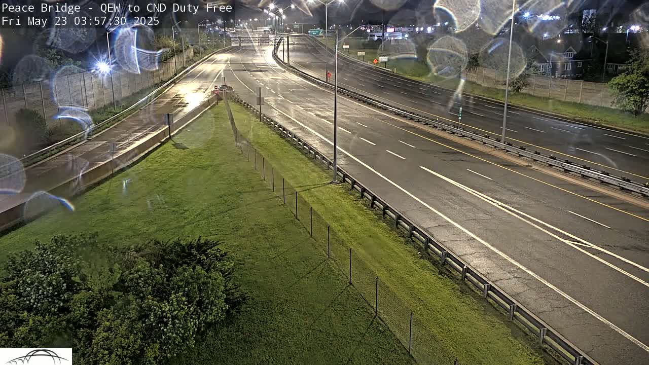 A mostly empty highway at night, wet from rain, curves gently to the left, bordered by a chain-link fence and grassy areas.
