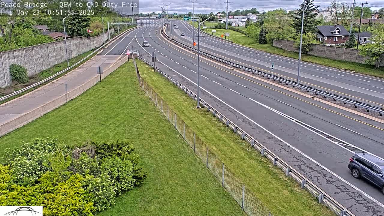 A multi-lane highway with sparse traffic on a partly cloudy day, bordered by grassy areas and chain-link fencing.