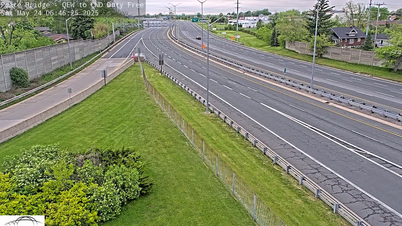A mostly empty multi-lane highway stretches into the distance under a partly cloudy sky, bordered by green grass and a chain-link fence.