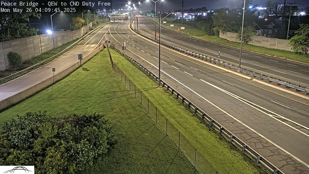 A mostly empty highway at night stretches into the distance, bordered by grassy areas and fences under clear skies.