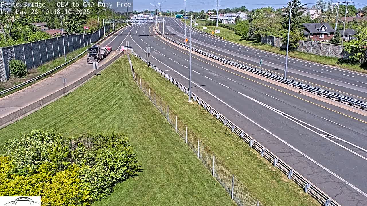 A multi-lane highway stretches into the distance under a sunny sky, with a grassy area and chain-link fence bordering one side, and a semi-truck visible in the near lane.