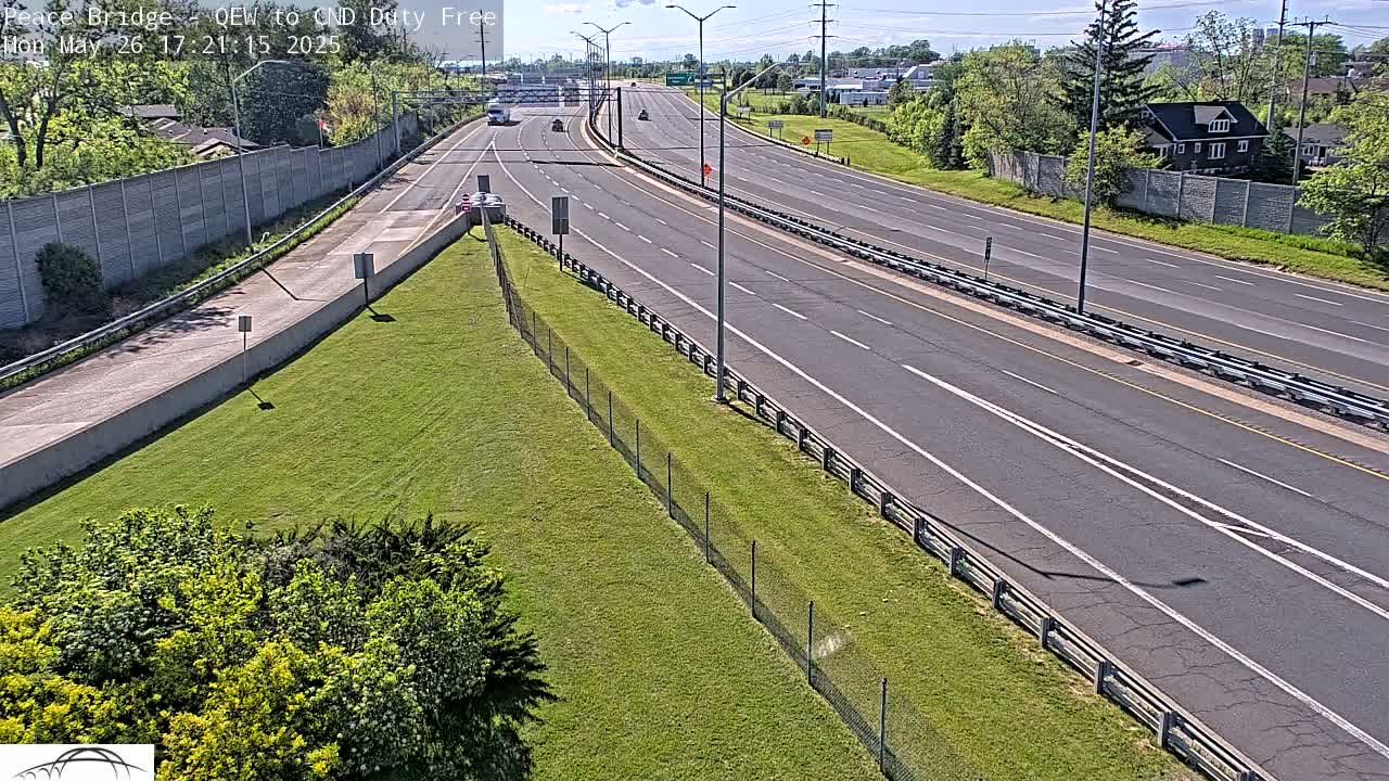 A multi-lane highway with sparse traffic on a sunny day, bordered by grassy areas and chain-link fencing.