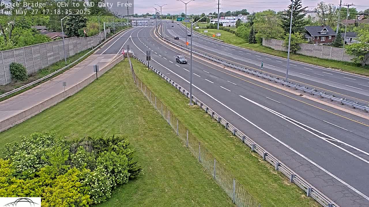 A multi-lane highway with sparse traffic under a partly cloudy sky, bordered by grassy areas and chain-link fences.