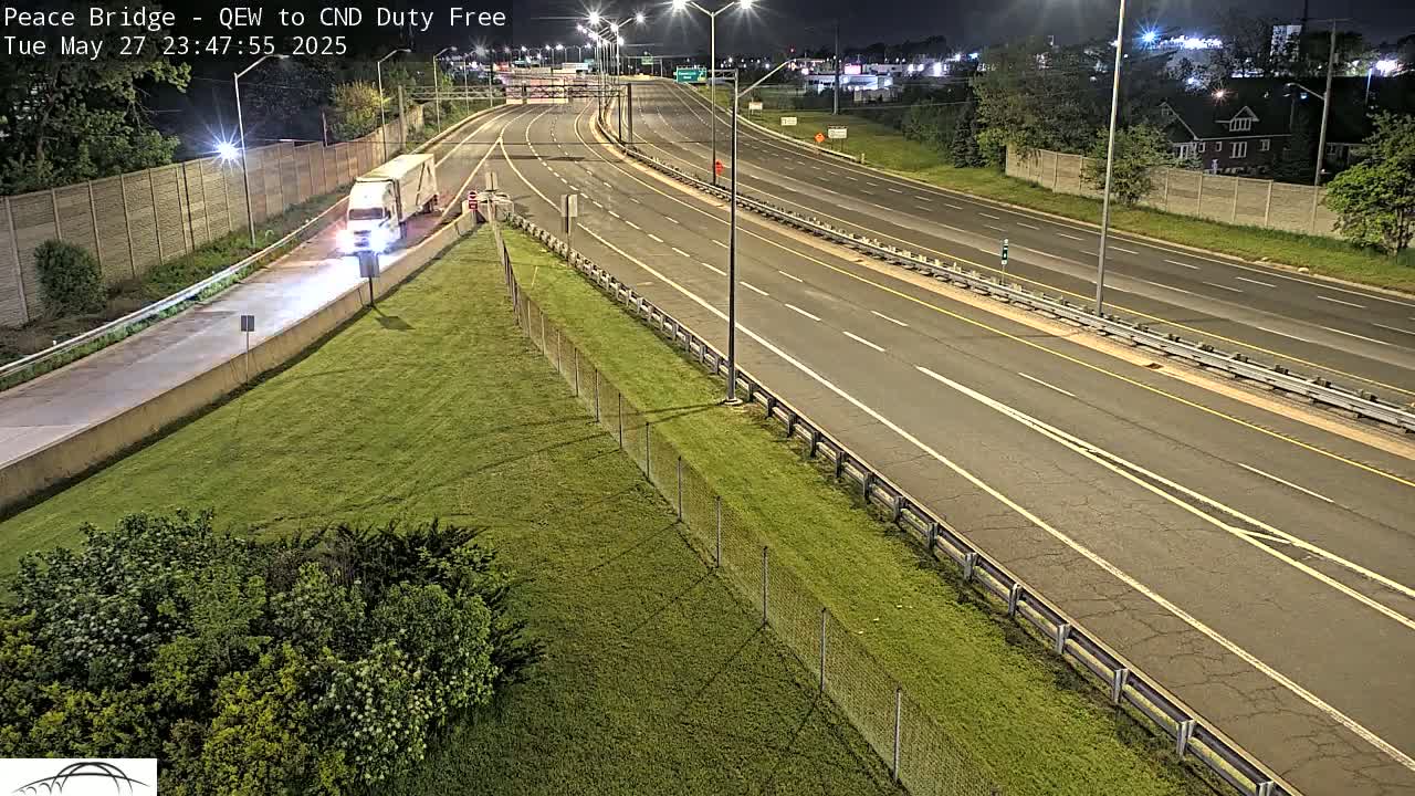 A white semi-truck is parked on the side of a multi-lane highway at night, with grassy areas and chain-link fencing visible.