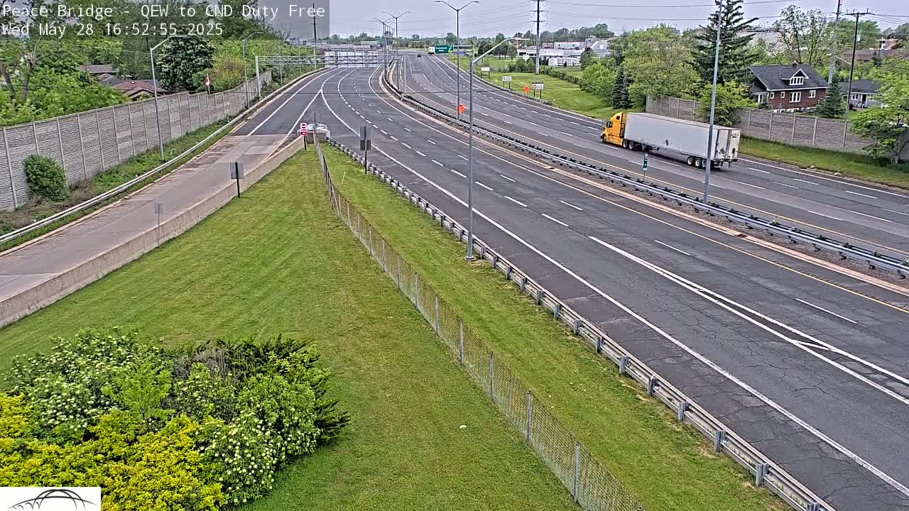 A multi-lane highway with sparse traffic on a partly cloudy day, bordered by grassy areas and a chain-link fence.