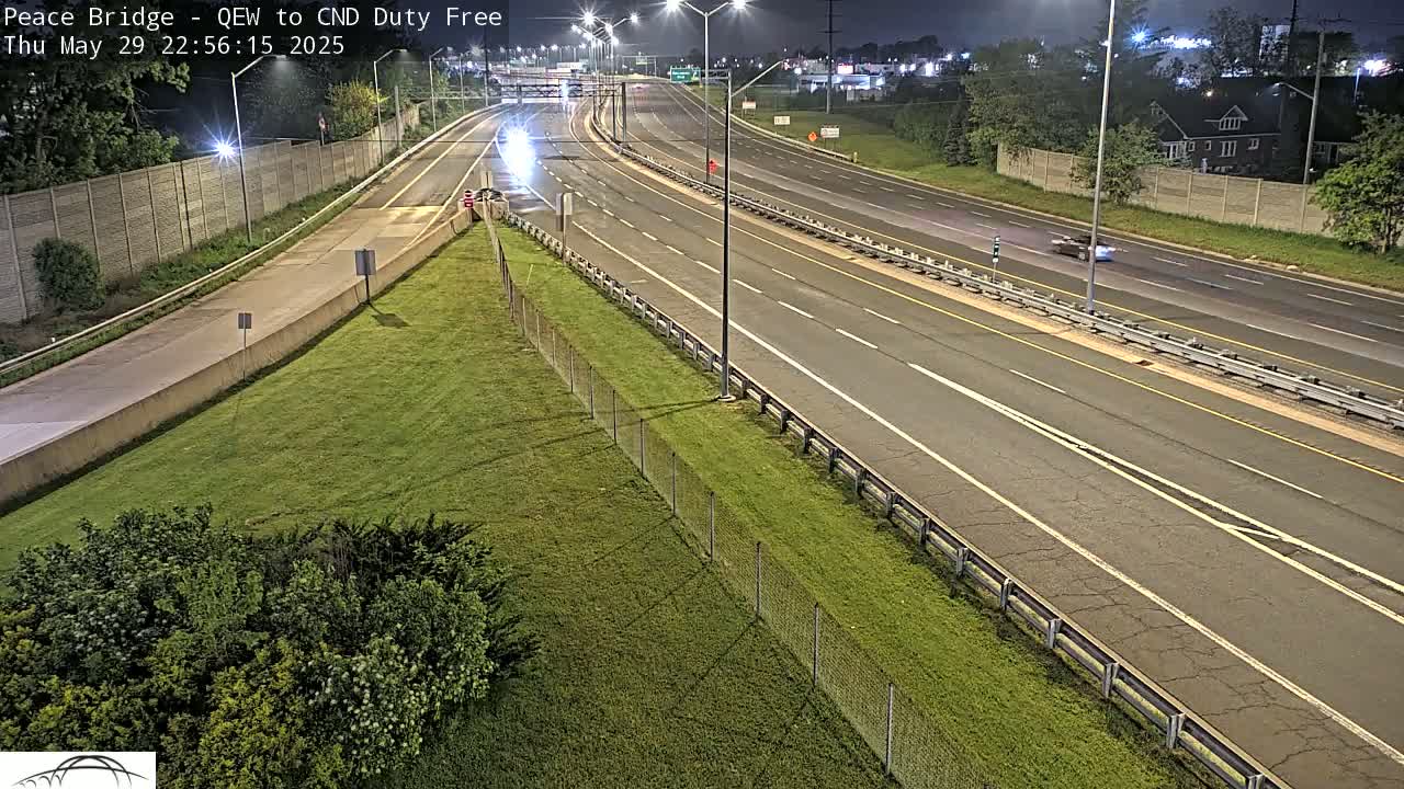 A nighttime, overhead view of a multi-lane highway with sparse traffic, bordered by grassy areas and fences.