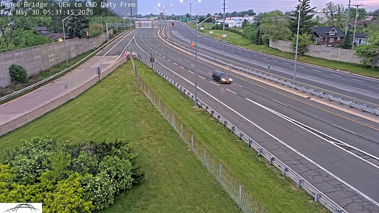 A multi-lane highway with sparse traffic is seen on an overcast day, bordered by grassy areas and a chain-link fence.