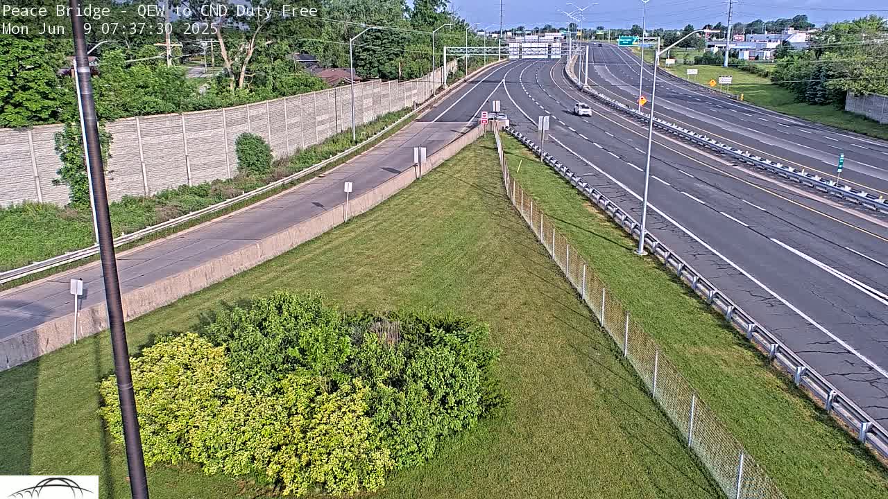 A high-angle view of a multi-lane highway on a sunny day, with sparse traffic, bordered by green grass and chain link fencing.
