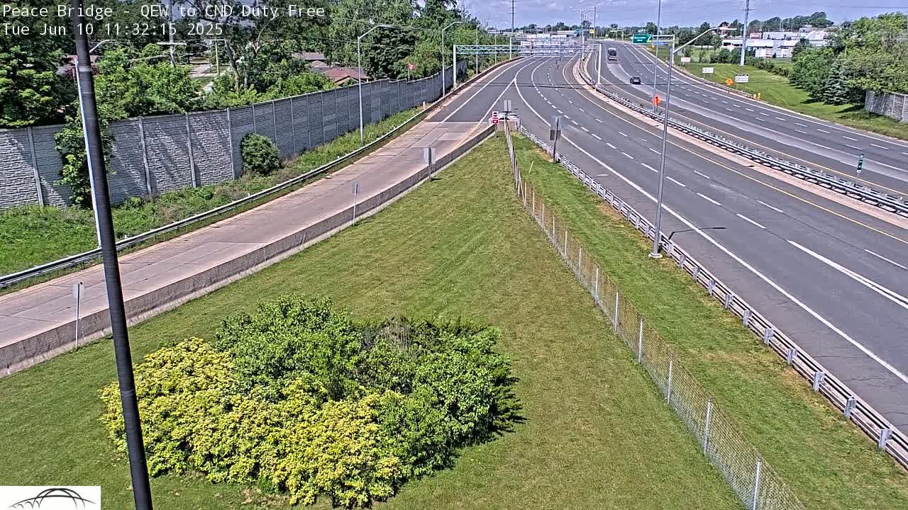 A highway scene on a sunny day shows a multi-lane highway with sparse traffic, bordered by grassy areas, chain-link fencing, and some shrubs.