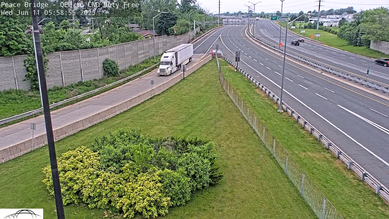 A white semi-truck travels on a multi-lane highway bordered by grassy areas and chain-link fencing under a partly sunny sky.