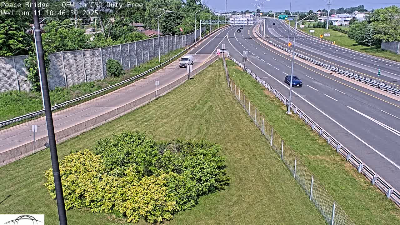 A high-angle view shows a highway on a sunny day with a few cars and a white van, surrounded by grassy areas and chain link fencing.