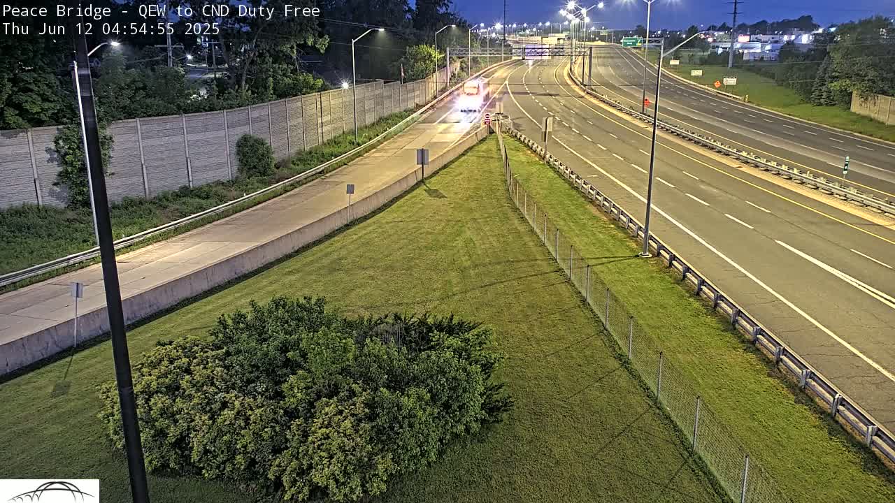 A nighttime overhead view of a multi-lane highway with sparse traffic, bordered by grassy areas and chain-link fencing.