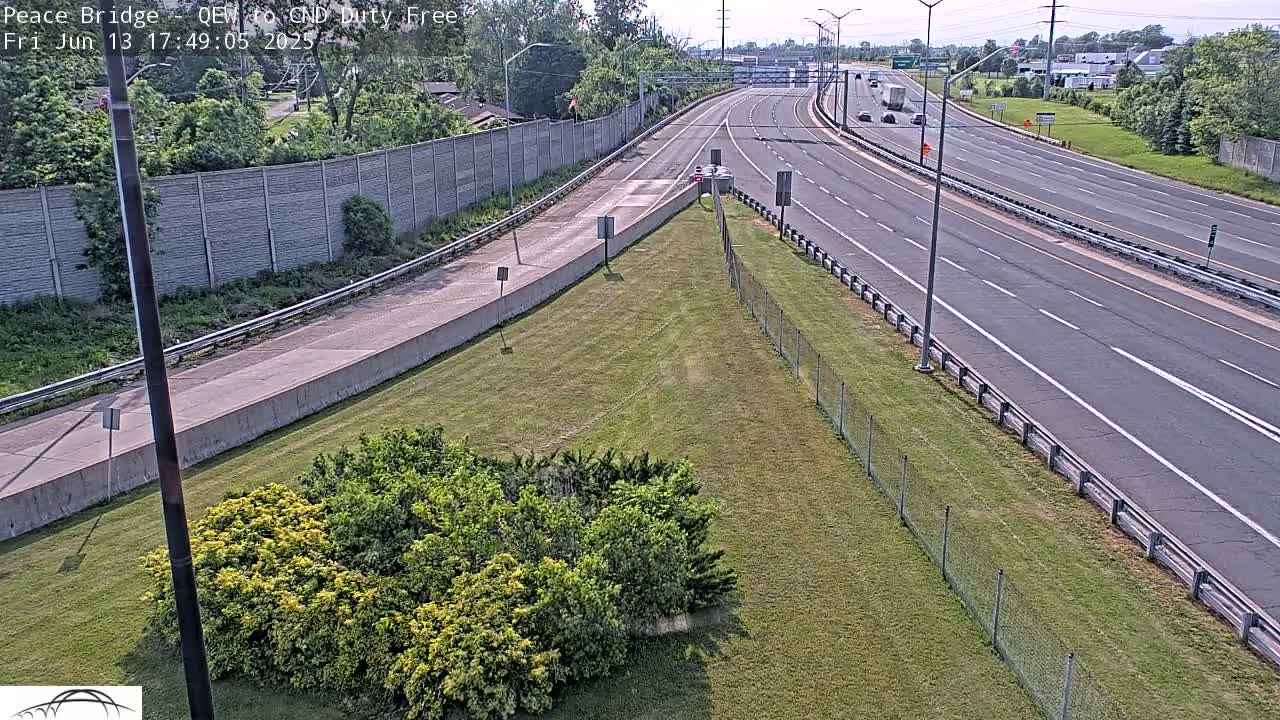 A highway with several vehicles on a sunny day, bordered by grassy areas and a tall concrete wall.