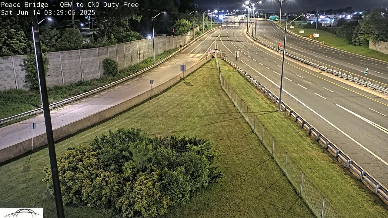 A high-angle nighttime view shows a multi-lane highway curving towards a grassy area bordered by a chain-link fence.