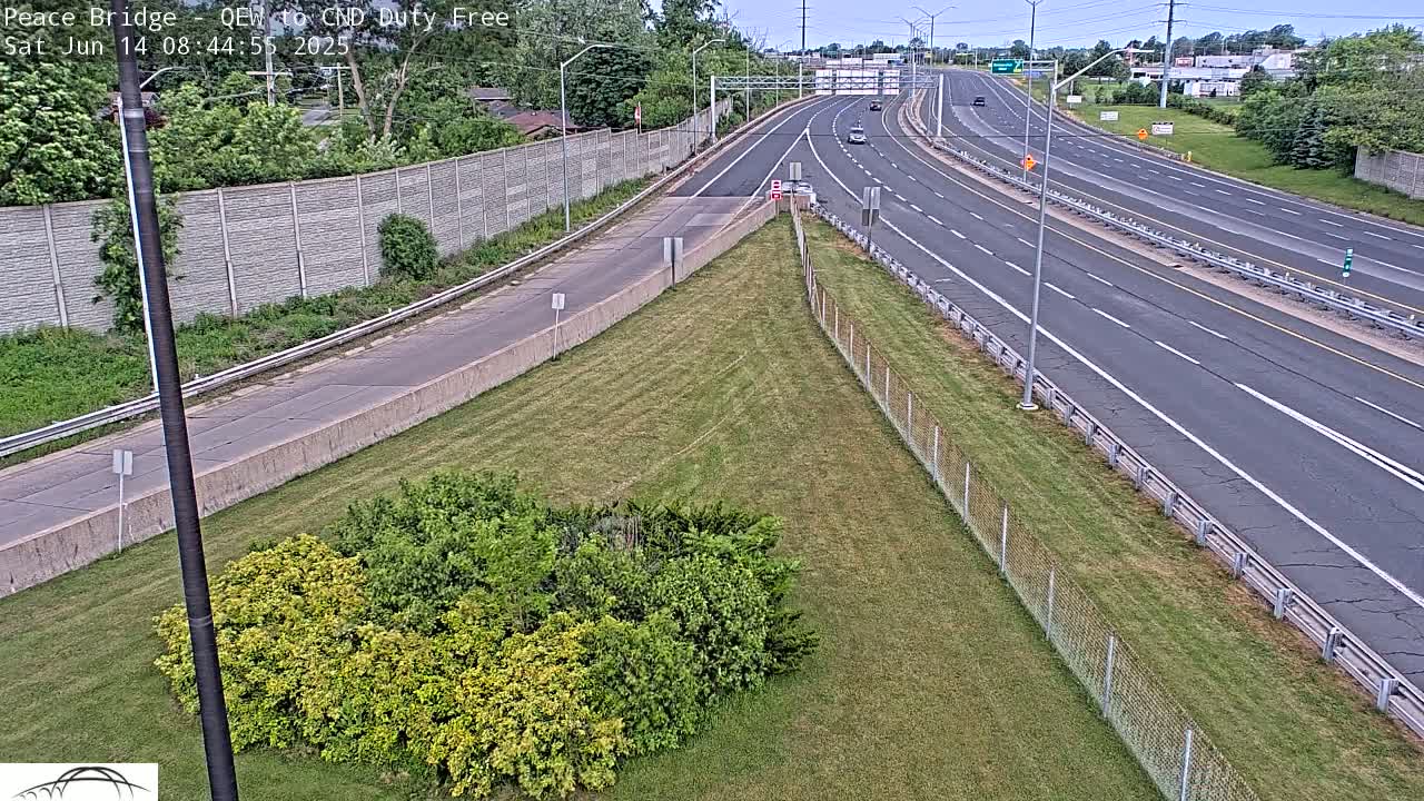 A mostly empty highway with a few cars under partly cloudy skies, bordered by a chain-link fence and grassy areas with some shrubs.